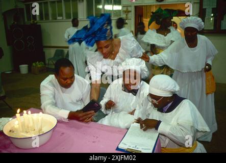 Baptême, cérémonie de baptême britannique noir, les membres de l'église viennent de danser autour de l'énorme pile de fruits. Il existe sept types de fruits et les bébés filles 8ème jour de la vie. Au milieu du groupe, un enfant est détenu par la Prophétesse, qui est en esprit. A sa gauche, un 'scribe' traduit les 'prophéties' et les écrit toutes. La Prophétesse parle en langues qui est l'un des dons surnaturels de l'esprit Saint mentionné dans 1 Corinthiens 12:4-10 : le père des bébés enregistre tout sur un magnétophone. Elephant and Castle, Londres, Angleterre, années 1993 1990 Royaume-Uni HOMER SYKES Banque D'Images