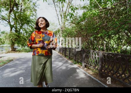 Femme asiatique avec un sourire joyeux et ouvert jouit de la tranquillité d'une journée d'été tout en marchant le long du trottoir du parc souriant femme d'âge moyen wa Banque D'Images