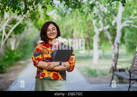 Une femme asiatique à la mode et souriante serrant un ordinateur portable se promène dans le parc. Personne branchée, douée de technologie, posant avec son appareil personnel à l'extérieur Banque D'Images