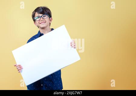 Garçon souriant joyeux avec des lunettes montrant la bannière blanche blanche en blanc pour la publicité d'affaires et regardant la caméra isolée sur fond jaune Banque D'Images