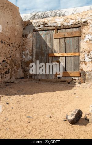 Très vieux bâtiment abandonné avec une porte en bois altérée. Sable dans la rue. Ciel bleu avec des nuages blancs. Caleta del Sebo, la Graciosa, Îles Canaries Banque D'Images