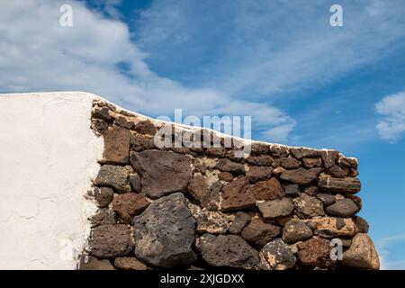 Mur fait de divers tons bruns de pierres volcaniques. Une autre partie avec une façade blanche. Ciel bleu avec des nuages blancs. Caleta del Sebo, la Graciosa, Canar Banque D'Images