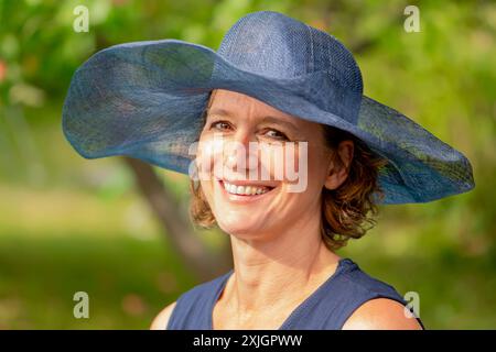 Une femme adulte aux cheveux bruns ondulés sourit tout en portant un chapeau bleu à larges bords. Banque D'Images