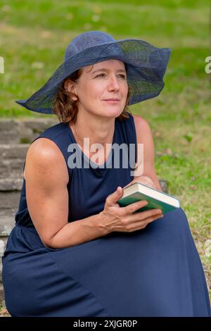 Une femme dans une robe bleue et un chapeau à larges bords est assise sur une marche de pierre dans un jardin herbeux, tenant un livre dans sa main. Banque D'Images