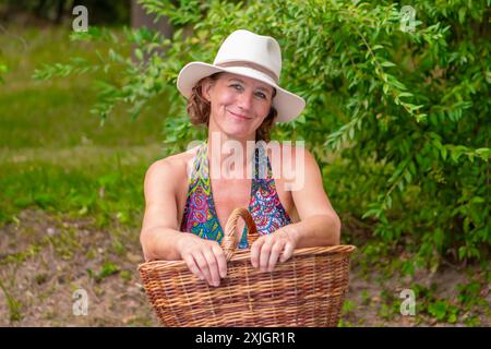 Une femme aux cheveux bruns courts et bouclés, portant un chapeau blanc et une robe fleurie colorée, tient un panier en osier devant elle. Elle est assise sur un banc Banque D'Images