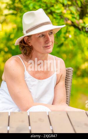 Une femme en robe blanche et chapeau blanc est assise à une table en plein air, la femme est entourée d'herbe verte et d'arbres. Banque D'Images