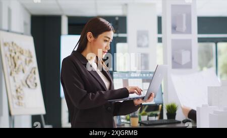 Développeur fait des esquisses sur ordinateur portable pour le programme de construction tout en inspectant le modèle de construction 3D sur la table de paramétrage dans l'espace de coworking. Entrepreneur développant le plan de rénovation et générant le plan. Banque D'Images