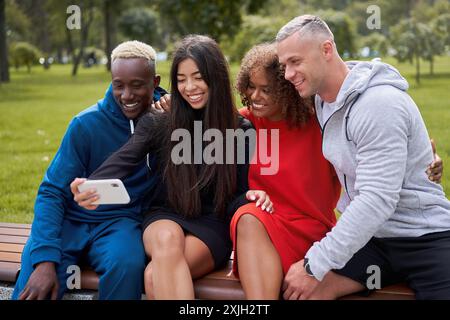 Souriant amis multi-ethniques prenant selfie sur smartphone. Des personnes de groupe diversifiées qui passent du temps ensemble assis sur un banc. Étudiants féminins masculins multiraciaux en vêtements de sport colorés pendant le week-end. Banque D'Images