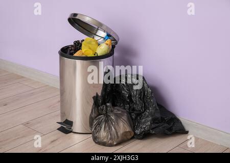 Poubelle métallique avec déchets alimentaires et sac poubelle plein près du mur lilas. Concept de recyclage Banque D'Images