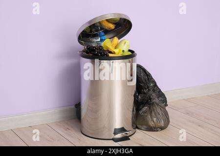 Poubelle métallique avec déchets alimentaires et sac poubelle plein près du mur lilas. Concept de recyclage Banque D'Images