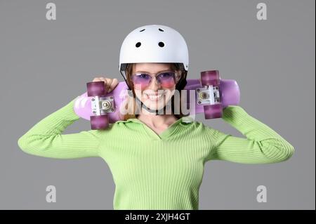 Belle jeune femme en casque avec skateboard sur fond gris Banque D'Images