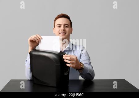 Beau jeune homme détruisant le papier dans la déchiqueteuse sur fond gris Banque D'Images