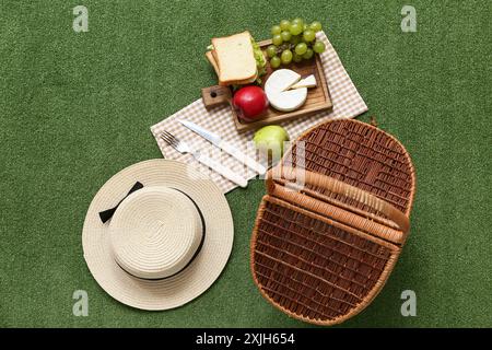 Panier de pique-nique en osier avec de la nourriture savoureuse et un chapeau élégant sur fond d'herbe verte Banque D'Images