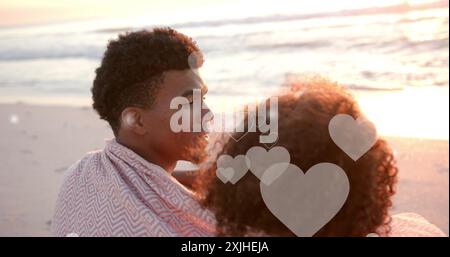 Image de cœurs se déplaçant sur un couple diversifié dans l'amour embrassant sur la plage en été Banque D'Images