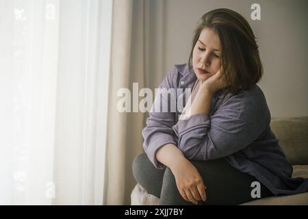 Femme de taille plus assise seule près de la fenêtre, portant des vêtements décontractés. Banque D'Images