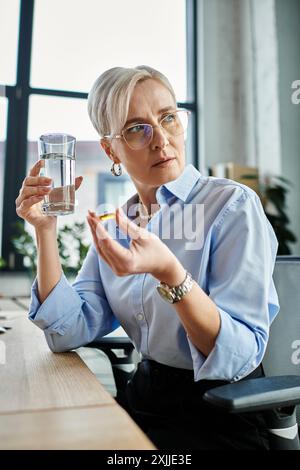 Une femme d'affaires concentrée d'âge moyen avec les cheveux courts gérant la ménopause, assise à un bureau tenant un verre d'eau. Banque D'Images