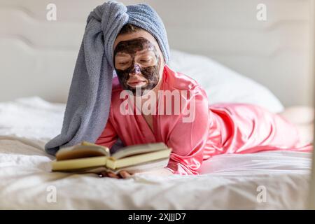 Femme avec masque noir et serviette sur la tête lisant livre sur le lit Banque D'Images
