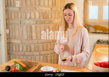 Belle fille blonde scooping avocat avec cuillère dans la cuisine Banque D'Images