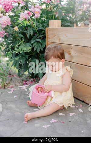 Mignon tout-petit en robe jaune assis avec arrosoir rose dans le jardin Banque D'Images