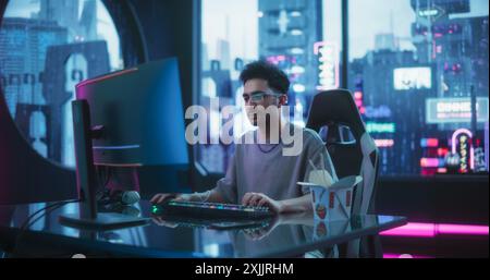 Beau jeune homme mangeant de la nourriture asiatique à emporter, tout en travaillant sur un ordinateur de bureau dans une salle Cyberpunk Neon. Pirate futuriste écrivant du code logiciel et portant des lunettes innovantes de réalité augmentée Banque D'Images