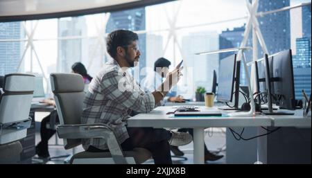 Portrait d'un beau manager indien utilisant un smartphone assis derrière un bureau dans un bureau avec un groupe de collègues. Souriant homme heureux répondant à un message, écrivant un billet en ligne drôle Banque D'Images