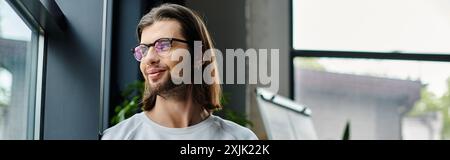 Bel homme caucasien avec de longs cheveux et des lunettes debout devant une fenêtre. Banque D'Images