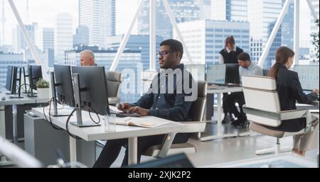 Analyste de données masculin afro-américain ciblé tapant sur ordinateur de bureau dans divers bureaux d'entreprise avec Megapolis Window View. Professionnel Balck homme travaillant dans la société de conseil avec des collègues. Banque D'Images