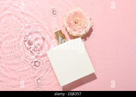 Belle fleur de rose et bouteille de parfum dans l'eau sur fond rose, pose plate Banque D'Images