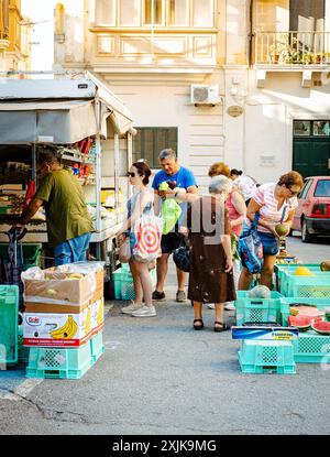 Un groupe diversifié de personnes faisant du shopping dans un marché agricole en plein air, parcourant des produits frais exposés dans des caisses, par une journée ensoleillée. Banque D'Images