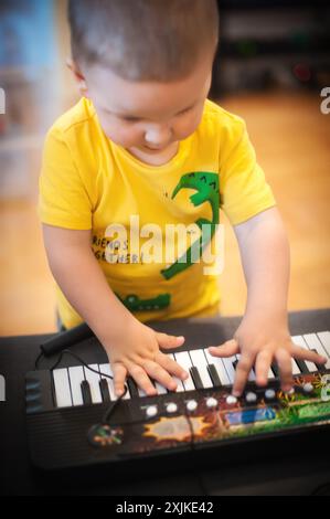 Heureux petit garçon est assis à un synthétiseur, ses doigts dansant sur les touches. Ses yeux sont focalisés sur l'instrument, son visage un mélange de concentration et Banque D'Images