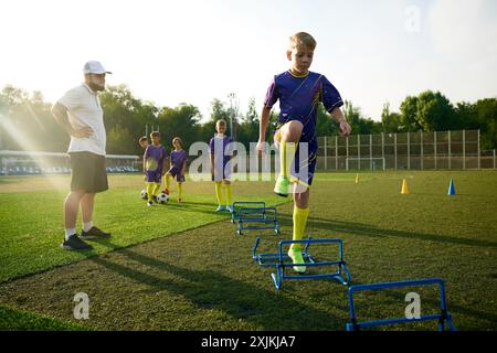 Garçons en uniforme, enfants pratiquant, exercices d'échauffement de pâte sur le sport déposé, sauter par-dessus les obstacles avant le match de football. Observation ciblée de l'entraîneur Banque D'Images