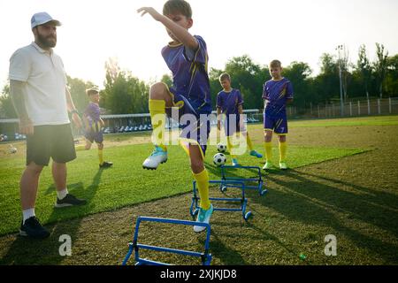 Garçons en uniforme, enfants pratiquant, exercices d'échauffement de pâte sur le sport déposé, sauter par-dessus les obstacles avant le match de football. Observation ciblée de l'entraîneur Banque D'Images