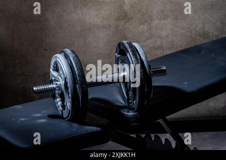 Gros plan d'un haltère en métal sur un banc d'entraînement noir dans une salle de sport faiblement éclairée, avec un arrière-plan flou Banque D'Images