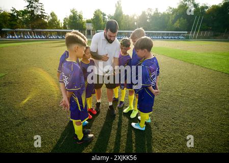 Garçons, enfants dans des uniformes vibrants se rassemblant autour de l'homme, entraîneur et écoutant la stratégie de jeu à l'aide d'une tablette. Stade extérieur Banque D'Images