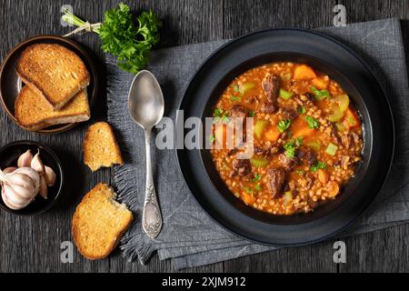 soupe d'orge de bœuf avec de tendres morceaux de rôti de bœuf, légumes frais et bouillon assaisonné dans un bol noir sur une table en bois de chêne foncé avec du pain et une cuillère, horiz Banque D'Images