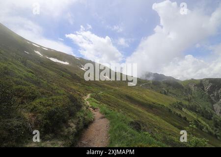 Vue sur une pente de montagne verte, à travers laquelle mène un étroit sentier de montagne et derrière laquelle des nuages blancs s'élèvent dans les airs Banque D'Images