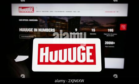 Stuttgart, Allemagne, 03-25-2023, personne tenant un téléphone portable avec le logo de la société polonaise de jeux en ligne HUUGE Inc. sur l'écran devant la page Web de l'entreprise. Banque D'Images