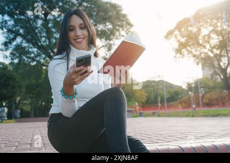 Heureuse jeune femme lisant les messages de son petit ami au téléphone, elle sourit joyeusement, assise dans le parc avec le coucher du soleil en arrière-plan Banque D'Images