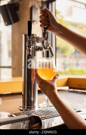 Gros plan des mains délicates d'une femme versant de la bière fraîche du robinet dans un verre dans un cadre de pub confortable. L'image capture la précision et le soin en soi Banque D'Images