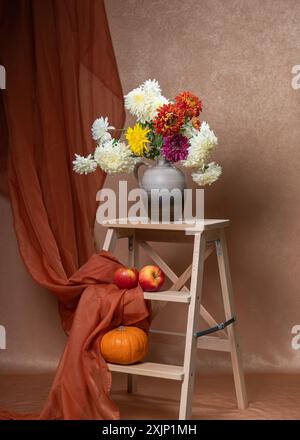 Un grand bouquet de fleurs d'automne blanches, jaunes, rouges. Beaucoup d'asters dans un vase gris sur un escalier en bois. Ci-dessous se trouvent deux pommes rouges, une petite citrouille. Ou Banque D'Images