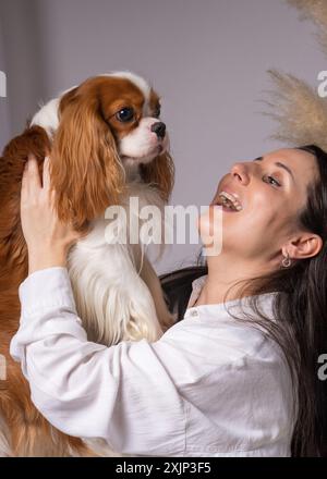 jeune femme enceinte assise sur le canapé à la maison. Riant joyeusement, il joue avec son chien. Cavalier roi charles, épagneul cocker Banque D'Images