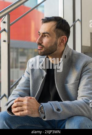 Portrait d'un beau jeune homme. Homme asiatique souriant avec une barbe soignée se trouve sur les marches près de la fenêtre et regarde au loin Banque D'Images