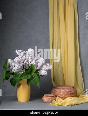 Un grand bouquet de lilas blancs dans un vase jaune sur un fond gris, à côté duquel est accroché un châle jaune et deux paniers ronds. Placer pour le texte. Banque D'Images