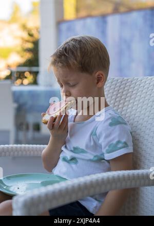 Petit déjeuner avant la maternelle. Mignon petit garçon mange un sandwich avec des saucisses, veut vraiment dormir, assis les yeux fermés et mange Banque D'Images