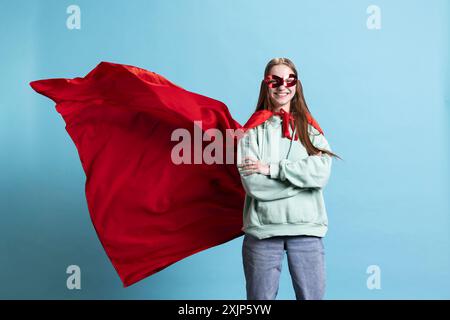 Jeune fille portant costume de super-héros et masque pour Halloween, bras croisés, fond de studio. Portrait d'une femme habillée en héros de bande dessinée cachant l'identité, debout fier Banque D'Images