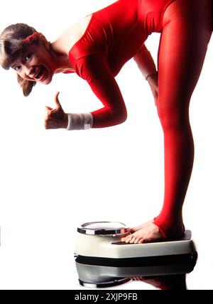 Une femme vêtue d'un léotard rouge se tient sur une balance avec ses pieds sur la plate-forme. Elle se penche en avant et regarde directement la caméra avec un bri Banque D'Images