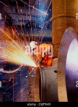 Un ouvrier dans une combinaison orange vif et un casque blanc utilise un outil de meulage sur un grand tuyau métallique à l'intérieur d'une usine. Le tuyau fait partie du con Banque D'Images