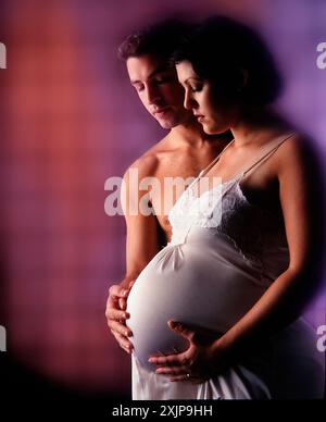 Une femme enceinte dans une robe blanche se tient avec son mari, qui lui tient le ventre. Le couple est debout devant un mur avec rouge et purpl muet Banque D'Images