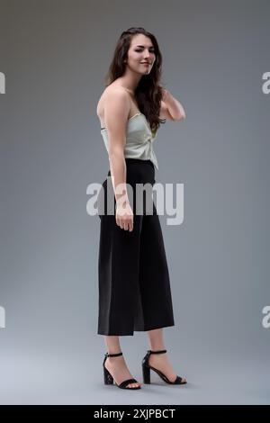 La jeune femme est debout dans une pose détendue, portant un pantalon noir taille haute et un haut blanc. Sa main repose sur ses cheveux Banque D'Images