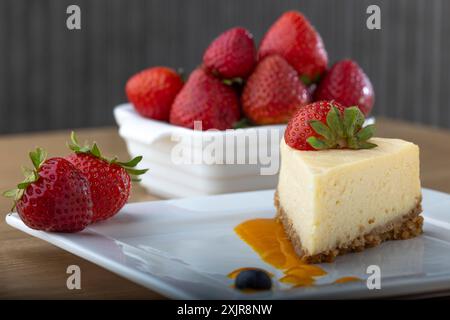 Une tranche de cheesecake crémeux garnie d’une fraise fraîche, servie sur une assiette blanche avec un filet de sauce aux fruits. Un bol de fraises fraîches Banque D'Images